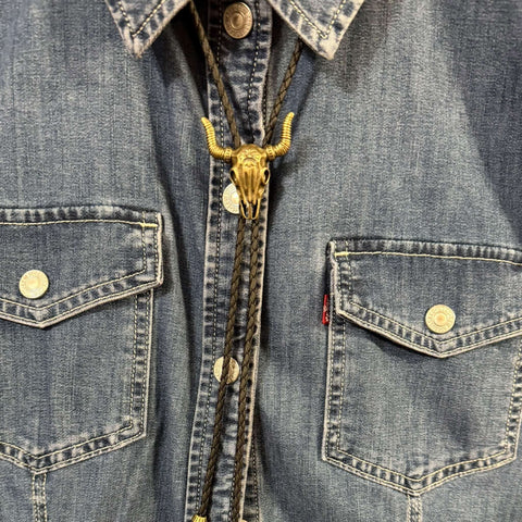 Antique gold tone steer skull bolo tie with black braided faux leather cord, adjustable slider pendant, and matching antique gold tips.