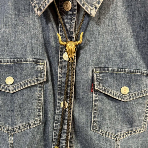 Antique gold steer skull bolo tie with adjustable slide and braided faux-leather rope. Unisex Western neckwear for concerts, festivals, and everyday edge.